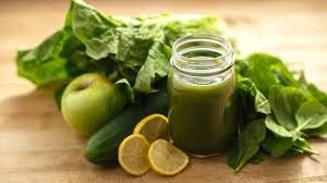 A small jar of green juice next to a cucumber, an apple, lemon, and salad leaves